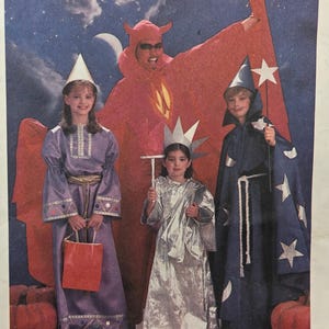 Puede incluir: Niños con disfraces de Halloween: un diablo rojo, un mago azul y una dama plateada. El diablo lleva cuernos y gafas de sol. La capa del mago está decorada con estrellas. Hay calabazas en primer plano.