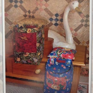 May include: A decorative arrangement featuring a white swan figurine, two fabric storage containers, and a folded fabric item. The containers have floral patterns and red accents. The background includes a patterned quilt.