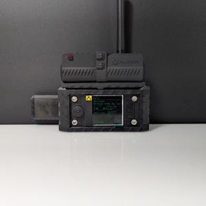May include: A black electronic device with a screen displaying green text and graphics. The device has a rectangular shape with a textured surface and is labeled "MILTECH". An antenna extends from the top. The device is on a white surface.