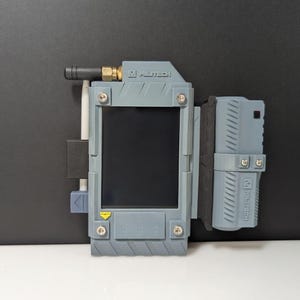 May include: A gray electronic device with a screen, antenna, and attached component. The device has the text "MILTECH" printed on it. The device is mounted on a black surface.