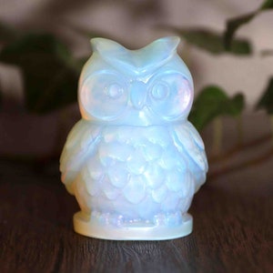 Könnte beinhalten: Eine weiße Opal-Eulenfigur mit schillernden Details. Die Eule sitzt auf einer Holzoberfläche.