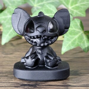May include: Black figurine of Stitch from Lilo & Stitch sitting on a black base. Stitch is a blue alien with large ears, two eyes, and a wide grin.