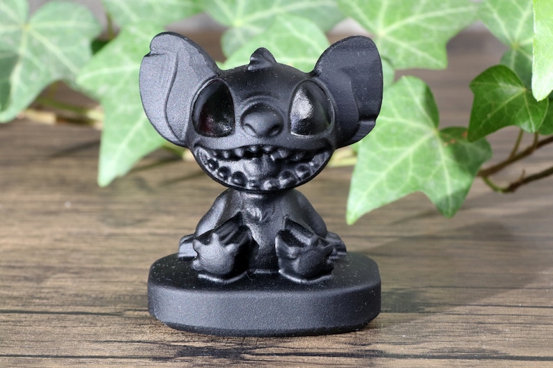 May include: Black figurine of Stitch from Lilo & Stitch sitting on a black base. Stitch is a blue alien with large ears, two eyes, and a wide grin.
