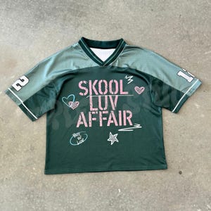 Puede incluir: Una camiseta de malla verde oscuro con mangas cortas y cuello en V. El frente presenta el texto rosa SKOOL LUV AFFAIR, junto con gráficos de corazones y estrellas en blanco y azul. El número 12 está impreso en blanco en ambas mangas.