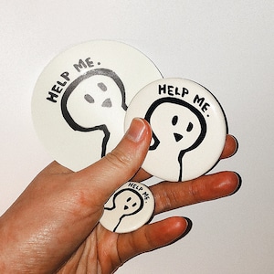 May include: Three white round pinback buttons with black outlines and the text "Help Me." Each button features a simple black and white illustration of a ghost-like figure.