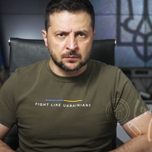 May include: A man wearing a green t-shirt with the text "Fight Like Ukrainians" in white and yellow. He is sitting in front of a blue and yellow flag and a screen with the Ukrainian coat of arms.