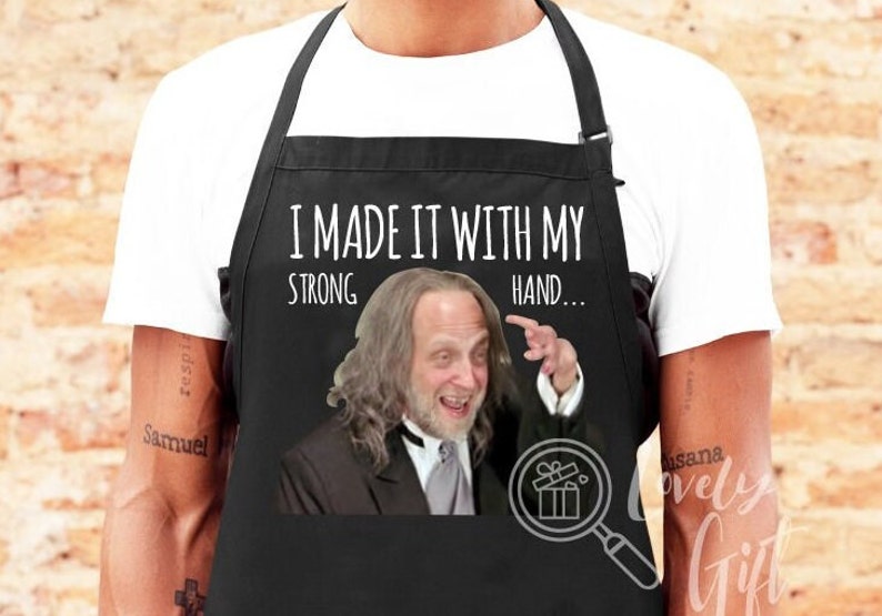 May include: Black apron with white text that reads "I made it with my strong hand..." and a photo of a man with long hair and a beard.