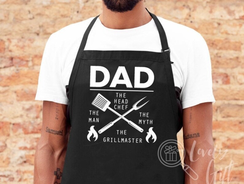 May include: Black apron with white text that reads "DAD THE HEAD CHEF THE MAN THE MYTH THE GRILLMASTER" with a spatula and grill fork crossed over the text.