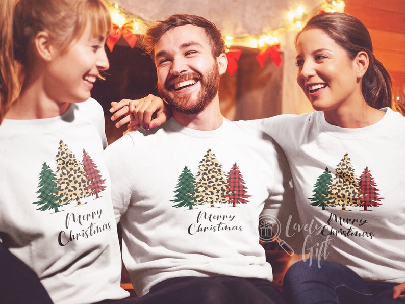 May include: Three people wearing white long-sleeved shirts with a Christmas tree design. The trees are green, red, and leopard print. The text "Merry Christmas" is printed on the shirts.