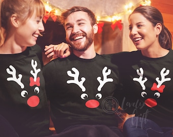 May include: Three people wearing black long-sleeved shirts with a white reindeer design. The reindeer has a red nose and a red bow. The people are smiling and looking at each other.