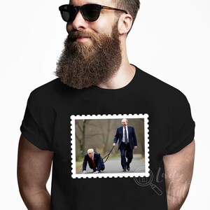 May include: A black t-shirt with a white border around a picture of two men. The man in the front is on his hands and knees, while the man behind him is walking and holding a leash attached to the man in front. The text "la" is visible in the bottom right corner of the image.