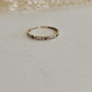 Könnte beinhalten: Ein goldener Bandring mit abwechselnd roten und klaren Edelsteinen. Der Ring liegt auf einer weißen Oberfläche mit einem verschwommenen Hintergrund aus getrockneten Pflanzen.