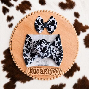 May include: A black and white cow print fabric hair bow and a matching knotted fabric headband. The headband is displayed on a white card featuring the text Little Birdtique and the URL littlebirdtique.etsy.com, with a wooden sign reading Little Birdtique.