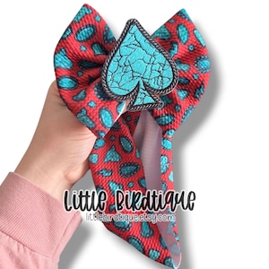May include: A red and turquoise paisley patterned hair bow with a turquoise spade-shaped embellishment. The bow features the text "Little Birdtique" and the Etsy shop URL.