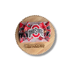 May include: A red and white Ohio State University bow on a wooden display. The bow features the Ohio State logo and other graphics. The wooden display has the text "Little Birdtique" on it.