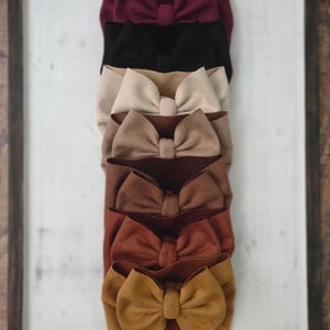 May include: A vertical stack of seven fabric headbands, each featuring a large, centered bow. The colors range from deep burgundy at the top, followed by black, cream, tan, brown, rust, and mustard yellow at the bottom. A wooden sign reads Little Birdtique.