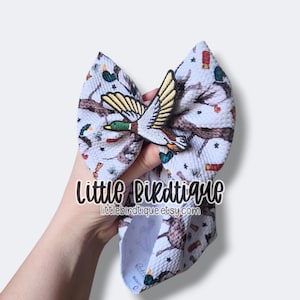 May include: A large, light-colored hair bow with a repeating pattern of ducks, shotguns, and tree branches. The bow features a 3D embroidered duck applique. The text "Little Birdtique" and the Etsy shop URL are visible.