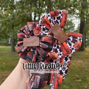 May include: A handmade hair bow featuring a brown and orange Wrangler print fabric with a cowhide pattern. The bow has a leather tag with the Wrangler logo. The text "Little Birdtique" and the Etsy shop URL are also visible.