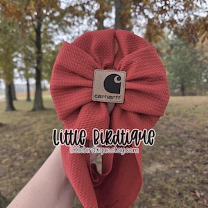 May include: A red baby bow with a Carhartt logo patch. The bow is made of textured fabric and has a large, decorative knot. The text "Little Birdtique" and the Etsy shop URL are visible.