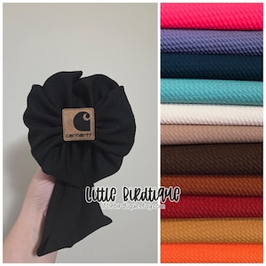 May include: Black baby bow with a Carhartt logo patch. The bow is made of textured fabric and has a large, flower-like shape. A variety of fabric colors are displayed in the background.