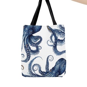 May include: A white tote bag featuring a watercolor design of blue octopuses. The bag has black handles and is held up by a person. The octopus design is repeated across the surface of the bag.