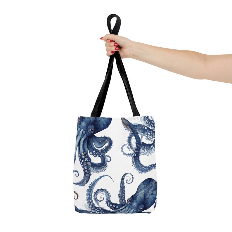 May include: A white tote bag featuring a watercolor design of blue octopuses. The bag has black handles and is held up by a person. The octopus design is repeated across the surface of the bag.