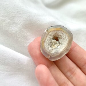 May include: A small, round, gray agate geode with a white crystal interior. The geode is about the size of a quarter and has a smooth, polished exterior.