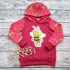 Könnte beinhalten: Ein magentafarbenes Kapuzensweatshirt mit einer Cartoon-Biene auf der Vorderseite. Die Ärmel und die Kapuze haben ein rosa Wabenmuster mit Bienenillustrationen. Das Wort "Lieblingsmensch" wird in Holzbuchstaben angezeigt.