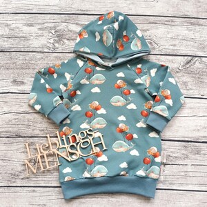 Könnte beinhalten: Türkisfarbener Kapuzenpullover mit einem verspielten Muster aus Walen, roten Luftballons und Wolken. Der Pullover hat eine Kapuze, lange Ärmel und gerippte Bündchen und Saum. Holzbuchstaben "Lieblingsmensch".