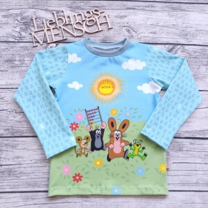 Könnte beinhalten: Ein hellblaues Langarmshirt mit einem grünen, gelben und blauen Druck von einer Sonne, Wolken, Blumen und Comicfiguren. Die Figuren beinhalten einen Maulwurf, ein Kaninchen und einen Frosch. Die Ärmel haben einen hellblauen Hintergrund mit einem Muster aus dem Alphabet in Grau.