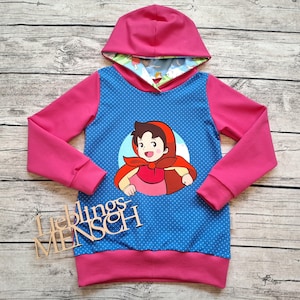 Könnte beinhalten: Ein rosa-blauer Kapuzenpullover mit einer Cartoonfigur in einem roten Umhang. Der Pullover hat rosa Ärmel und einen blauen Körper mit weißen Punkten. Das Kapuzenfutter hat ein farbenfrohes Muster. Das Wort "Lieblingsmensch" ist sichtbar.