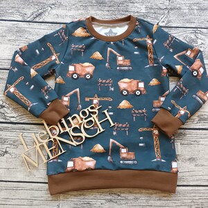 Könnte beinhalten: Ein tealblauer Langarm-Sweatshirt mit braunem geripptem Kragen und Bündchen. Der Sweatshirt hat ein sich wiederholendes Muster aus Baumaschinen, darunter Muldenkipper, Bagger und Kräne. Der Text "110" ist am Kragen sichtbar.