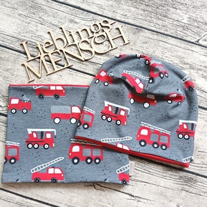Könnte beinhalten: Eine graue und rote Strickmütze und ein passender Halsschmuck mit einem Feuerwehrauto-Muster. Mütze und Halsschmuck sind aus weichem, dehnbarem Stoff.