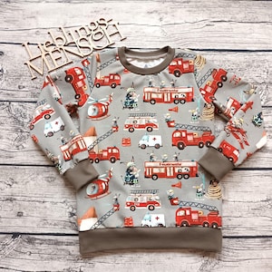 Könnte beinhalten: Ein graues Langarmshirt mit Rundhalsausschnitt und einem Druck von Feuerwehrautos, Hubschraubern und anderen feuerbezogenen Bildern. Das Shirt hat einen braunen gerippten Bund und Saum.