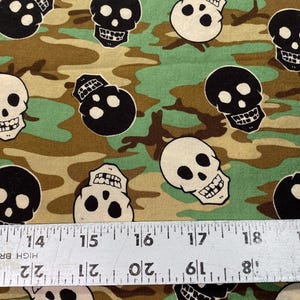 Op de afbeelding: Een stof met een camouflagepatroon met zwarte en witte schedelontwerpen. De stof is bruin, groen en beige.