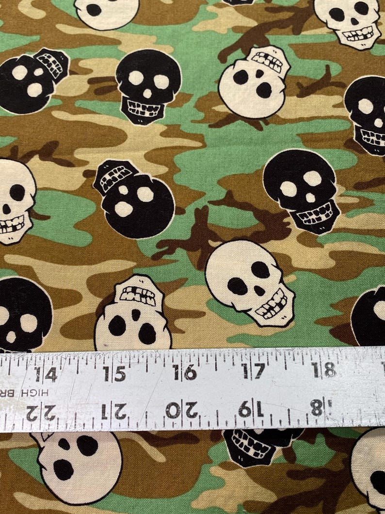 Op de afbeelding: Een stof met een camouflagepatroon met zwarte en witte schedelontwerpen. De stof is bruin, groen en beige.