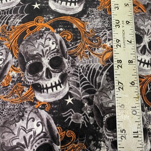 Op de afbeelding: Zwarte stof met een herhalend patroon van witte en grijze suikerschedels met oranje bloemmotieven. De stof heeft ook witte sterren en zwarte spinnen op een web.