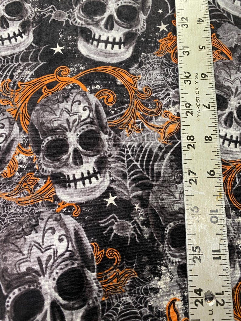 Op de afbeelding: Zwarte stof met een herhalend patroon van witte en grijze suikerschedels met oranje bloemmotieven. De stof heeft ook witte sterren en zwarte spinnen op een web.