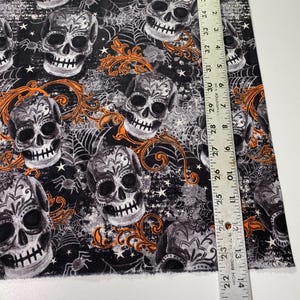 Op de afbeelding: Zwarte stof met een herhalend patroon van witte en grijze suikerschedels met oranje bloemmotieven en spinnenwebben. De stof wordt gemeten met een liniaal in inches.