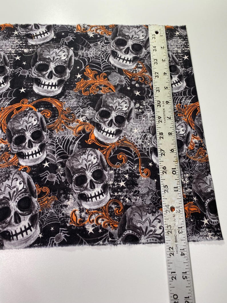 Op de afbeelding: Zwarte stof met een herhalend patroon van witte en grijze suikerschedels met oranje bloemmotieven en spinnenwebben. De stof wordt gemeten met een liniaal in inches.