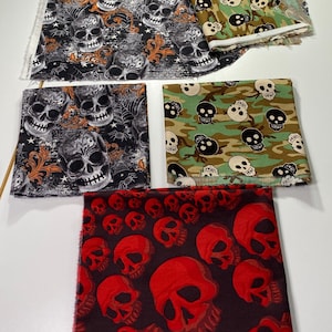 May include: Four different fabric swatches with skull patterns. The first swatch has a black background with white and orange skulls, the second has a black background with white and orange skulls, the third has a camouflage background with black and white skulls, and the fourth has a black background with red skulls.