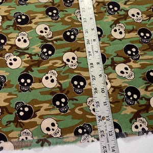 Op de afbeelding: Groen, bruin en zwart camouflage-stof met een herhalend patroon van witte en zwarte schedelontwerpen.