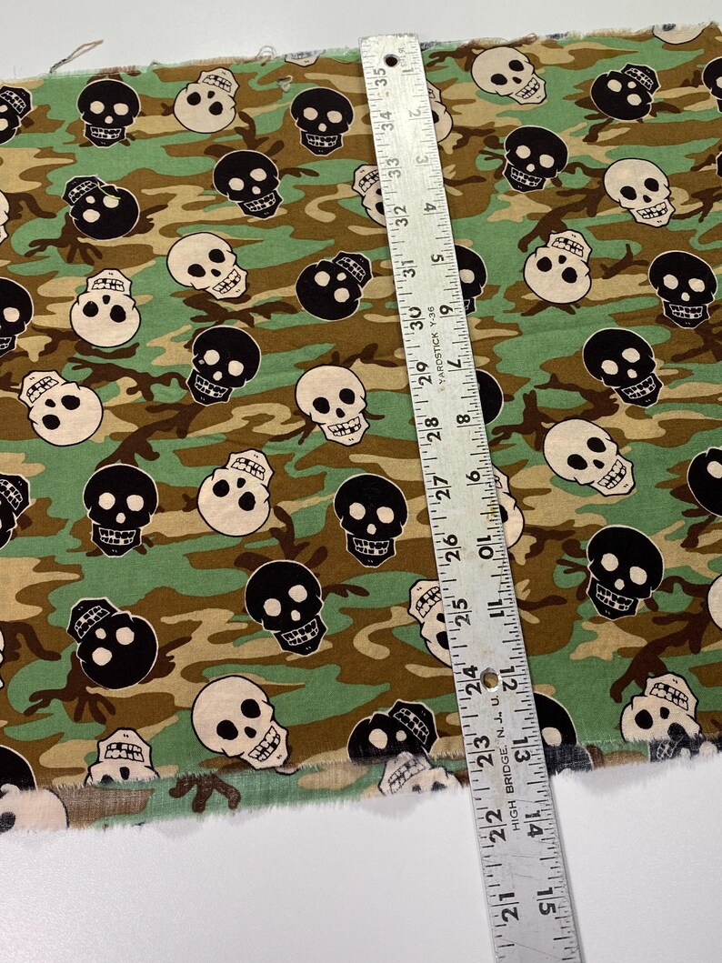 Op de afbeelding: Groen, bruin en zwart camouflage-stof met een herhalend patroon van witte en zwarte schedelontwerpen.