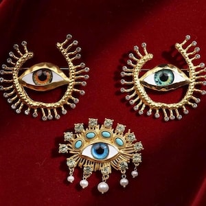 Può includere: Tre spille a forma di occhio color oro con disegni intricati. Ogni spilla presenta un occhio centrale con iride dettagliata e una cornice decorativa. Un occhio è marrone, uno verde e uno blu con accenti turchesi e perle pendenti.