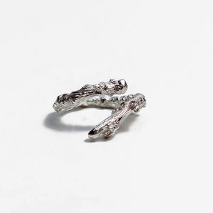 Può includere: Un anello d'argento a forma di due rami intrecciati. L'anello è realizzato in metallo testurizzato che ricorda la corteccia di un albero.