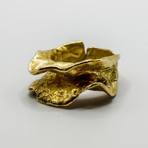 Puede incluir: Un anillo de color dorado con un diseño único, orgánico y texturizado. El anillo es ancho y tiene una forma ondulada e irregular.