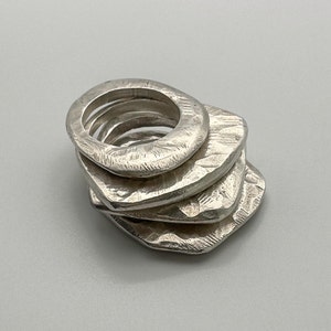 May include: A stack of five silver rings, each with a unique textured surface. The rings are stacked in a graduated size, with the largest ring at the bottom and the smallest ring at the top.