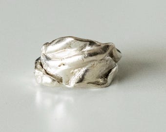 May include: A silver ring with an irregular, organic, and textured surface. The metal band features deep folds and ridges, creating a multi-layered appearance with a reflective, polished finish that catches the light across its uneven form.
