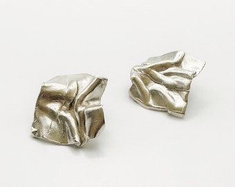 May include: A pair of silver-colored stud earrings. Each earring has a crumpled, textured appearance, with multiple folds and creases. The earrings are set against a plain white background, highlighting their metallic sheen and unique design.