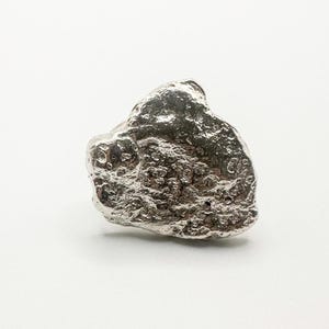 Può includere: Un anello argentato di forma irregolare con una superficie strutturata. L'anello ha un aspetto naturale e organico, simile a un piccolo sasso o pepita. L'anello è posto su uno sfondo bianco.